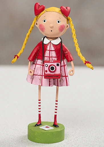 Toy figure of a girl with yellow pigtails, red heart-shaped hair accessories, and a pink camera, standing on a green base.  lori mitchell valentine's day new 2027 figurines