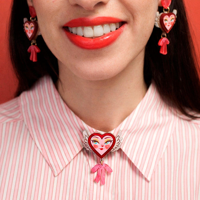 Person wearing a pink striped shirt with a heart-shaped brooch and matching earrings against a red background