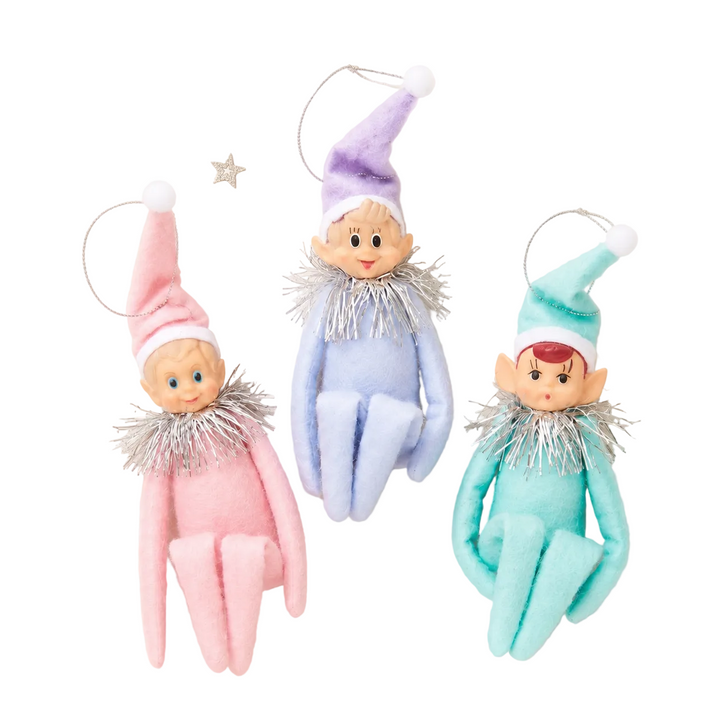 Three plush elf ornaments in pink, light blue, and mint green hang against a warm cream background with small silver star decorations. Each elf has a pointed hat, silver tinsel collar, and a metal hanging loop. kneeghugger elf