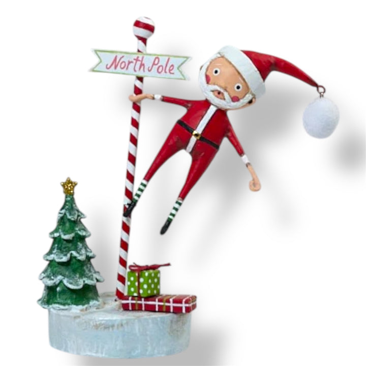 A festive figurine of Santa Claus holding a 'North Pole' sign, standing next to a Christmas tree and a present, designed by Lori Mitchell.