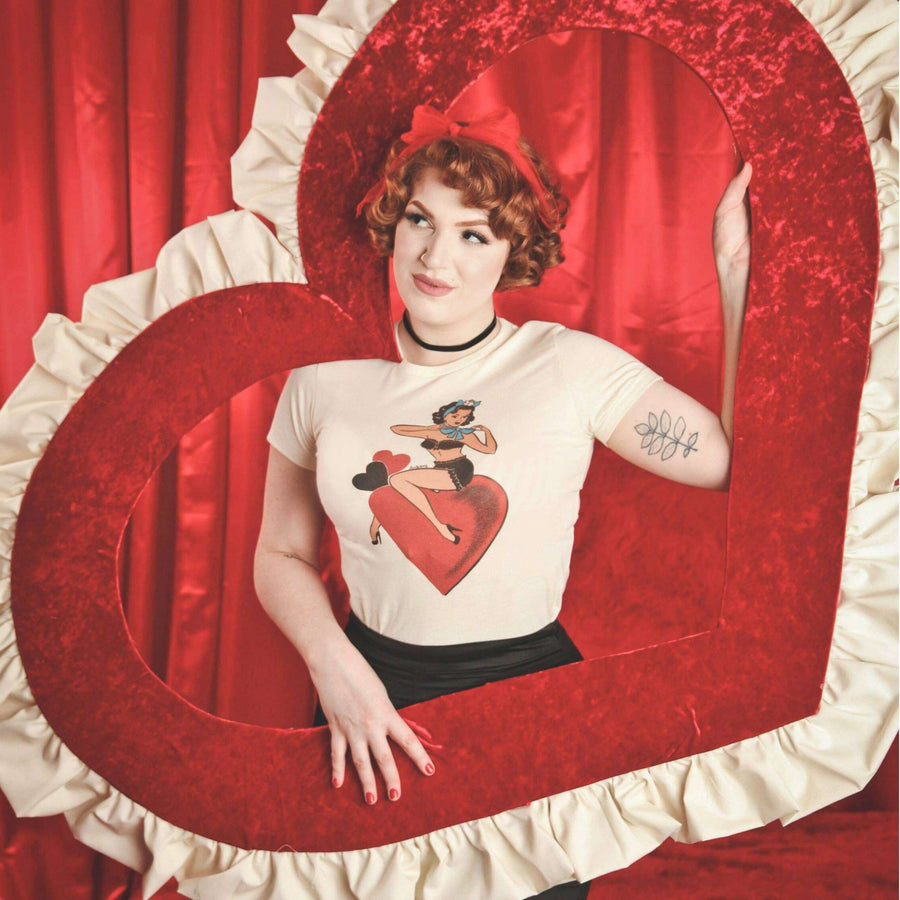 Person wearing a t-shirt with a graphic design in front of a heart-shaped mirror on a red curtain background