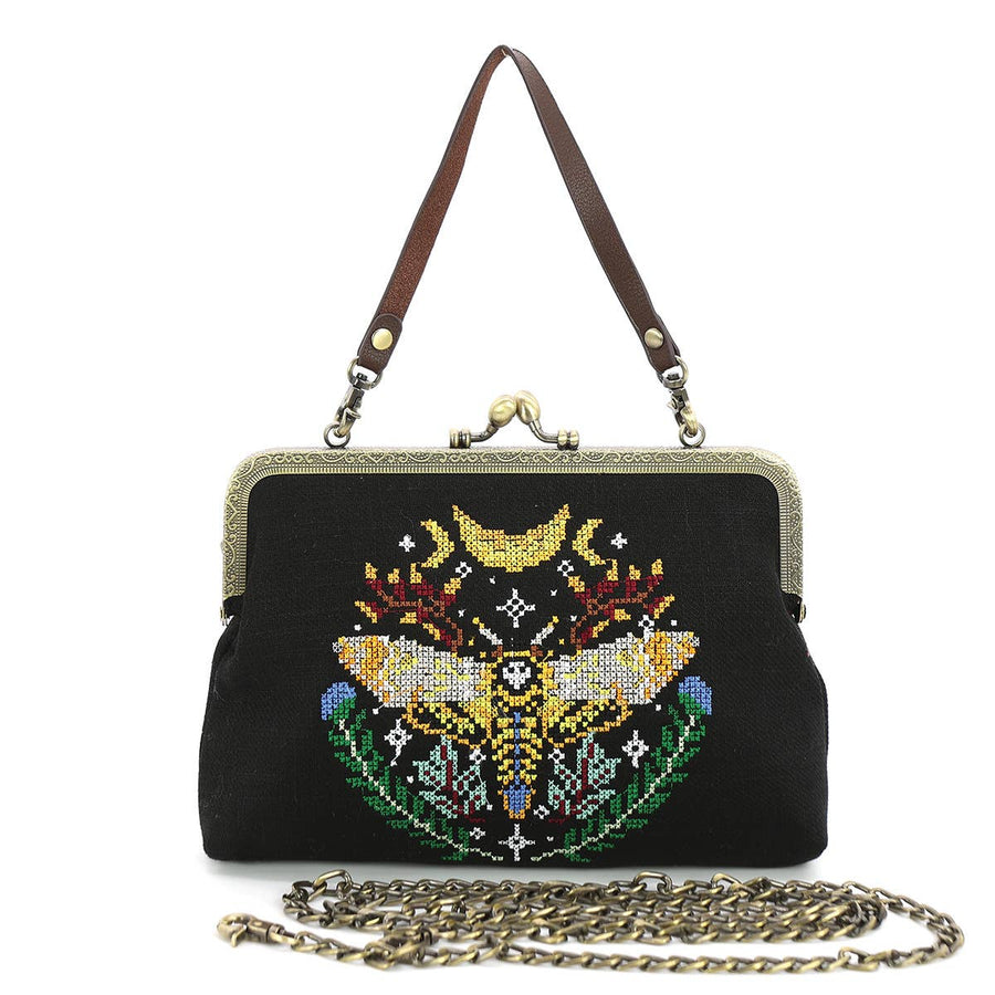 A vintage style handbag with a skull and moth embroidered design on a black canvas background, featuring a metal frame clasp and a leather strap.